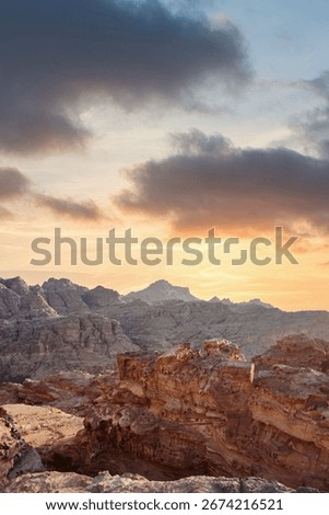 The sun sets behind rugged mountain peaks, casting a warm glow over the rocky landscape. Unique rock formations contrast against the colorful sky, creating a tranquil atmosphere. Petra, Jordan.