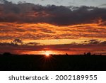 The sun sets behind shadowed fields  in Iowa.  Blue, yellow, orange and red clouds show brilliance with sun star at the horizon.  Light blue sky at the top of the image.