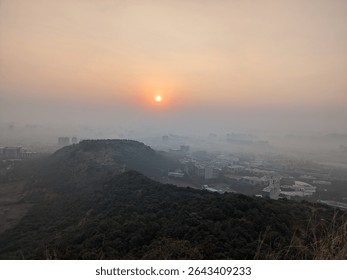 The sun sets behind sparse, leafless trees on a hilltop, casting a gentle glow over a foggy city below. A calm and serene end-of-day atmosphere.