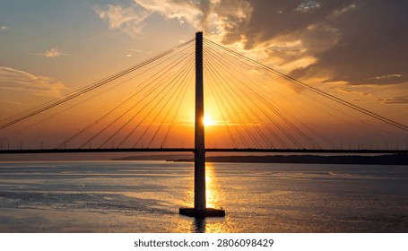 The sun sets behind a striking cable-stayed bridge, casting a warm glow over the water and sky.
