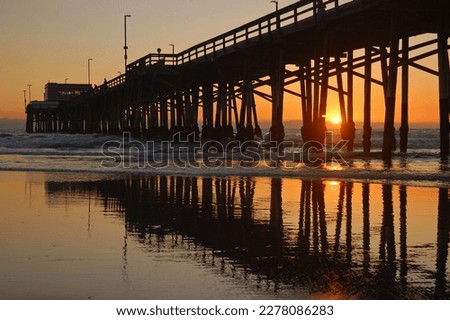 The sun sets between the pylons of the Newport Beach pier and is reflected in the wet sand of the shore