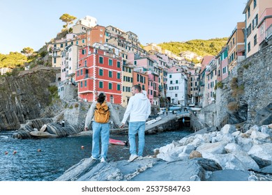 As the sun sets, a couple admires the vibrant buildings of Riomaggiore while standing on the rocky shore, soaking in the enchanting atmosphere of this charming Italian village. - Powered by Shutterstock