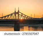 The sun sets directly behind a large metal bridge with ornate towers, casting a warm glow over the river