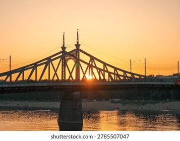 The sun sets directly behind a large metal bridge with ornate towers, casting a warm glow over the river