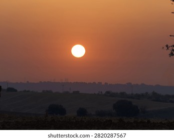 A sun sets in the evening sky over open fields. The horizon shows a mix of land and trees. The sky takes on orange hues as daylight fades.