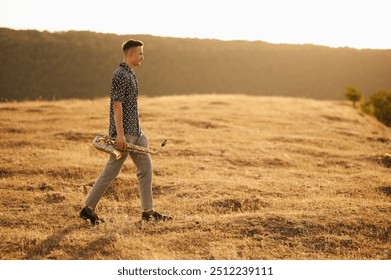 As the sun sets, a man strolls through a vast expansive field, holding onto a musical instrument