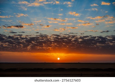 The sun sets over a flat desert landscape with dark foreground mounds and scattered clouds lit in orange and blue tones
