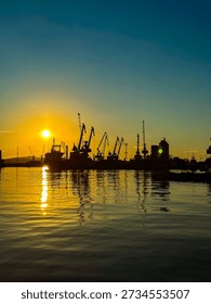 The sun sets over a harbor, creating a bright light that shines on the water. Silhouettes of cranes and ships are seen along the dock.