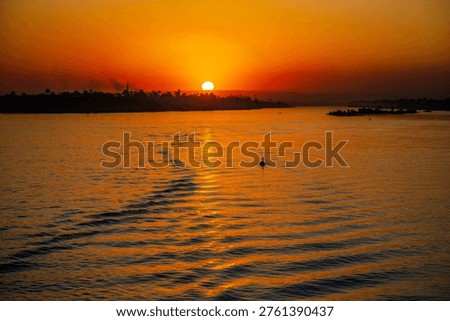 The sun sets over the Nile River near Luxor, casting orange light across the rippled water and distant riverbank