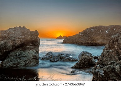 The sun sets over the ocean, casting a warm orange glow on the horizon. Waves gently lap against the rocky shore, creating a serene and tranquil atmosphere.