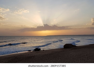 The sun sets over the ocean, casting light across the water. Waves gently hit the shore while clouds scatter above. It is early evening at a quiet beach.
