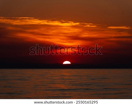 The sun sets over the ocean, casting bright orange and red hues across the sky. Calm water reflects these colors, creating a serene and tranquil atmosphere as day turns to night.