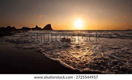 The sun sets over the ocean, casting a warm glow on the waves as they crash against the rocky shore. Hartland, Devon UK