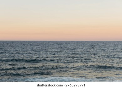 The sun sets over the ocean, creating a soft glow in the sky. The waves gently roll onto the shore. A distant horizon forms the boundary between sea and sky as colors blend.