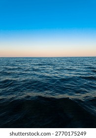 The sun sets over the ocean, creating a line of soft colors in the sky. Waves gently roll in. The scene shows a clear blue sky meeting the water at the horizon during the evening.