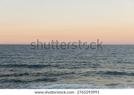 The sun sets over the ocean, creating a soft glow in the sky. The waves gently roll onto the shore. A distant horizon forms the boundary between sea and sky as colors blend.