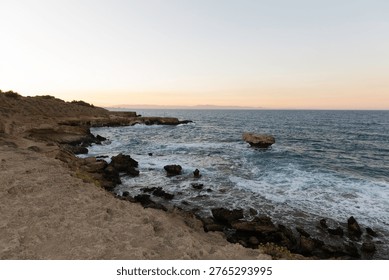 The sun sets over the ocean as waves crash on the rocky shore. The sky shows colors of orange and purple. Mountains are visible in the distance.
