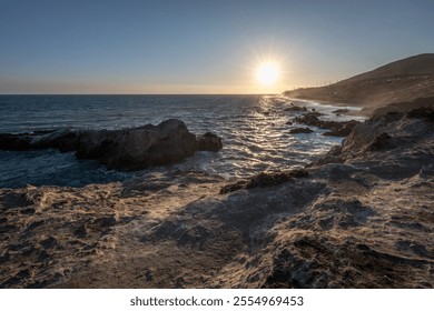 The sun sets over the Pacific Ocean at Leo Carrillo State Beach, casting golden light on the rocky shoreline and gentle waves. - Powered by Shutterstock