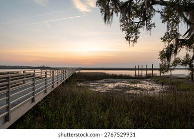 The sun sets over the river, casting light on a wooden pier. Grass grows along the water's edge. The sky shows colors of dusk. The scene captures the end of the day.