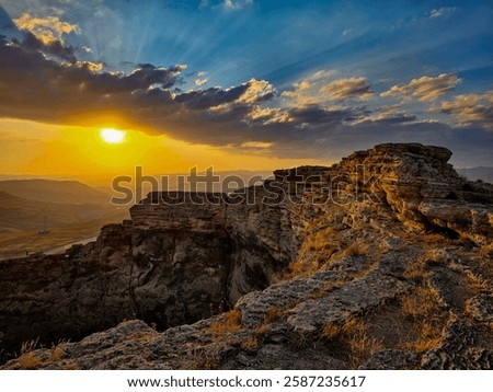As the sun sets over the rugged cliffs, golden light bathes the rocky formations, highlighting their intricate textures and weathered edges. The sky is a breathtaking blend of deep blues and fiery ora