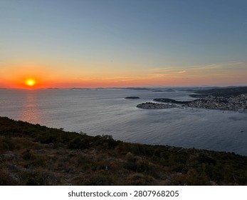 The sun sets over the water, casting light on the sea and islands. A small town with buildings is seen along the coastline. The sky shows colors of orange and pink during evening hours.