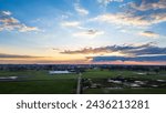 As the sun sets, its rays peek through cloud cover, casting a dramatic light over expansive farmland and facilities below. Sunset Sky Over Farmland: A Majestic Display of Clouds and Light. High