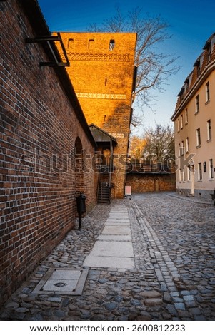 As the sun sets, warm light washes over the Leaning Tower in Torun, illuminating the cobblestone street and medieval architecture.