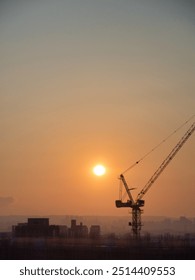 Sun setting behind a construction crane, illuminating the hazy skyline, casting long shadows across the urban landscape, creating a warm, industrial atmosphere.