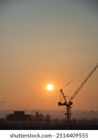 Sun setting behind a construction crane, illuminating the hazy skyline, casting long shadows across the urban landscape, creating a warm, industrial atmosphere.