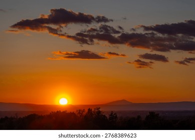 Sun setting behind distant mountains and silhouetted trees with orange sky and clouds