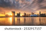 Sun setting behind a modern skyline, river reflecting golden hues, with dramatic clouds and a geometric blue building adding a futuristic touch.