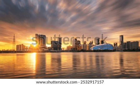 Sun setting behind a modern skyline, river reflecting golden hues, with dramatic clouds and a geometric blue building adding a futuristic touch.