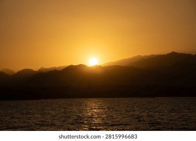 Sun setting behind mountain range, casting warm light over tranquil ocean water