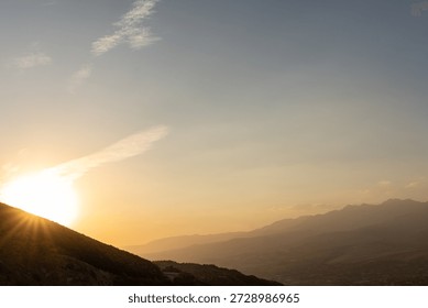 Sun is setting behind mountains as shadows grow over the landscape. A dam stands by the water while a winding road leads into the scene. The sky has warm colors.