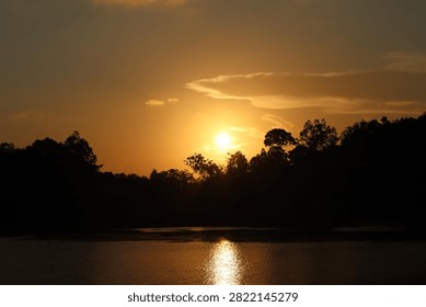 Sun setting behind tree line silhouette over lake water with golden light reflection.