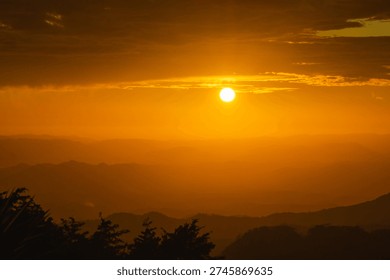 Sun setting, casting a warm orange glow over rolling mountains and distant trees