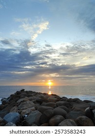 The sun setting in the middle of the ocean and rocks at the edge of the beach.