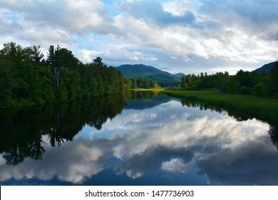 The sun is setting on a quiet, tranquil evening, making the Ausable River become mirror-like, reflecting the Adirondack Mountains in the background.