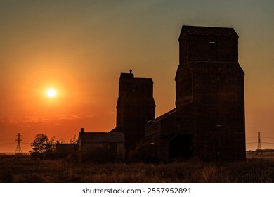 The sun is setting over a field with two old grain silos. The sky is a mix of orange and pink hues, creating a serene and peaceful atmosphere