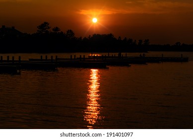 The sun setting over a lake with empty docks on the water.  Silhouetted trees are seen in against the orange sky. 