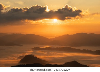 The sun is setting over a mountain range, casting a warm glow over the landscape. The sky is filled with clouds, creating a sense of mystery and depth. The mountains are covered in fog