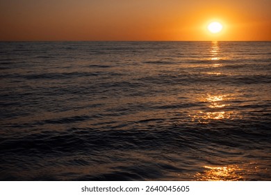 The sun is setting over the ocean, casting a warm glow on the water. The sky is a mix of orange and pink hues, creating a serene and peaceful atmosphere. The water is calm and still