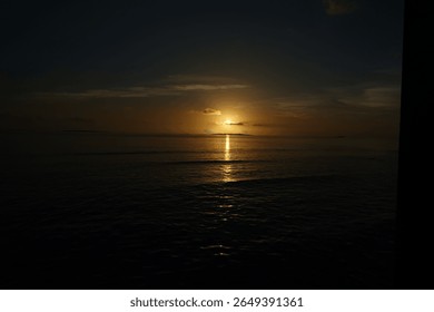 The sun is setting over the ocean, casting a warm glow on the water. The sky is darkening, and the water is calm. The scene is peaceful and serene