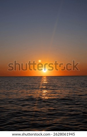 The sun is setting over the ocean, casting a warm glow on the water. The sky is a mix of orange and pink hues, creating a serene and peaceful atmosphere. The water is calm