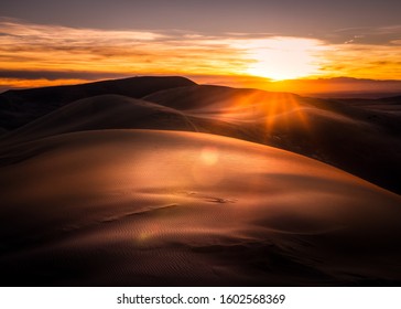 Sun setting over sand dunes in Colorado. The sky and sand is very orange. There are sun rays and a little lens flare.