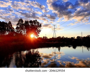 Sun Setting Over Suburban Wetlands Lake With Near Mirror Like Reflection Of Clouds And Blue Sky Off The Water, And Large Overland Powerlines.