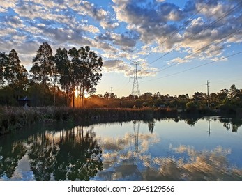 Sun Setting Over Suburban Wetlands Lake With Near Mirror Like Reflection Of Clouds And Blue Sky Off The Water, And Large Overland Powerlines.