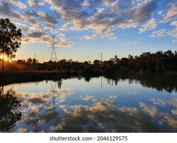 Sun Setting Over Suburban Wetlands Lake With Near Mirror Like Reflection Of Clouds And Blue Sky Off The Water, And Large Overland Powerlines.