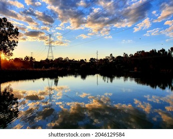 Sun setting over suburban wetlands lake with near mirror like reflection of clouds and blue sky off the water, and large overland powerlines.