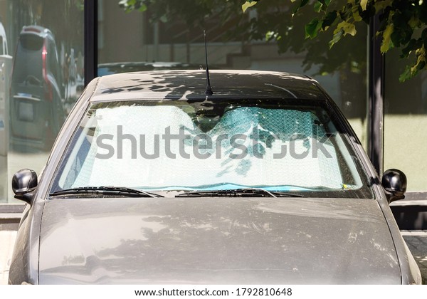 Sun shades\
under the windshield protects gray car from the sun while parked\
outdoorson a day. Foil sun shield made of metallic silver foil\
reflects direct sunlight. Front\
view.